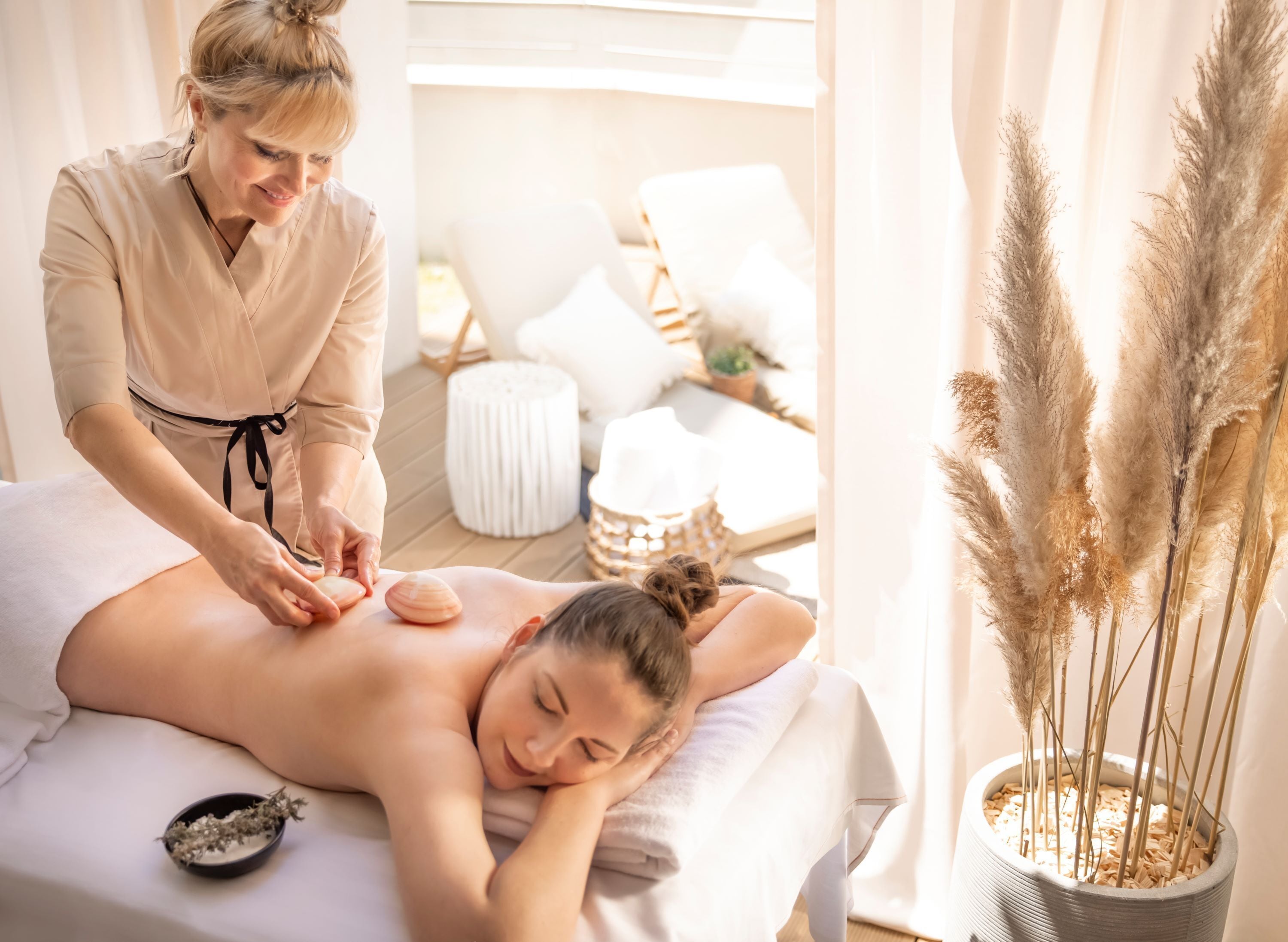 A massage therapy session in a well-lit room with a massage table, a bowl of dried herbs, and a potted plant with pampas grass.