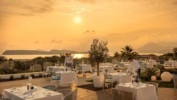 Un espace de restauration en plein air, avec des tables dressées pour les clients, offrant une vue panoramique sur l’océan et les montagnes au coucher du soleil.
