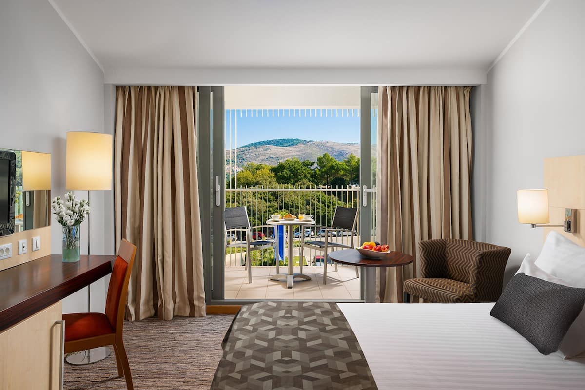 Habitación de hotel con cama, escritorio, silla, una pequeña mesa con otra silla, y un balcón con mesa y sillas.