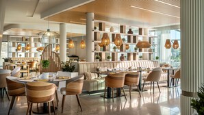Un restaurante moderno con techo alto, paneles de madera e iluminación colgante. Hay varias zonas de estar con mesas y sillas, y un área central con sofás.