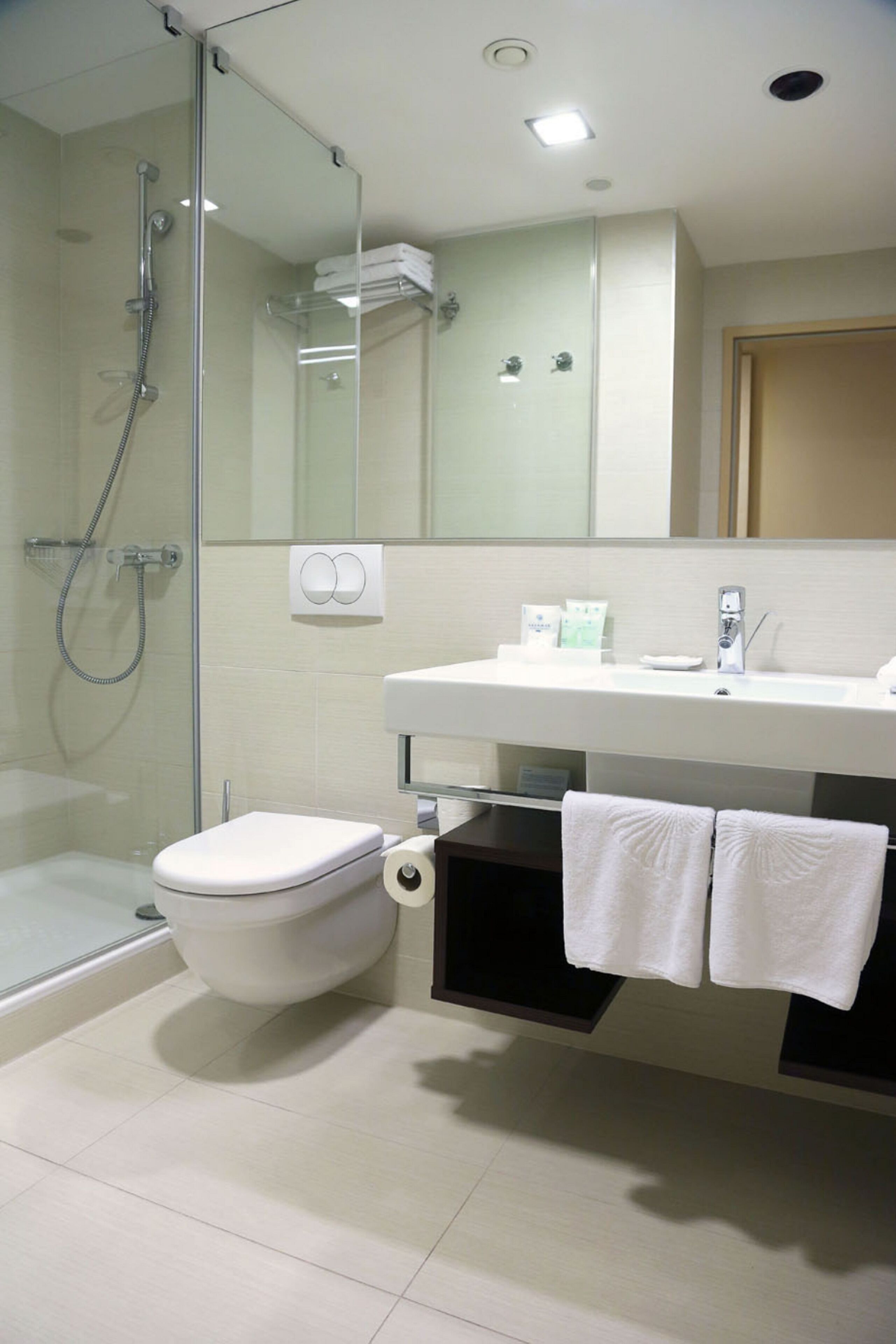 Un baño moderno con ducha de vidrio, inodoro blanco, lavabo con espejo y dos toallas colgadas en un perchero.