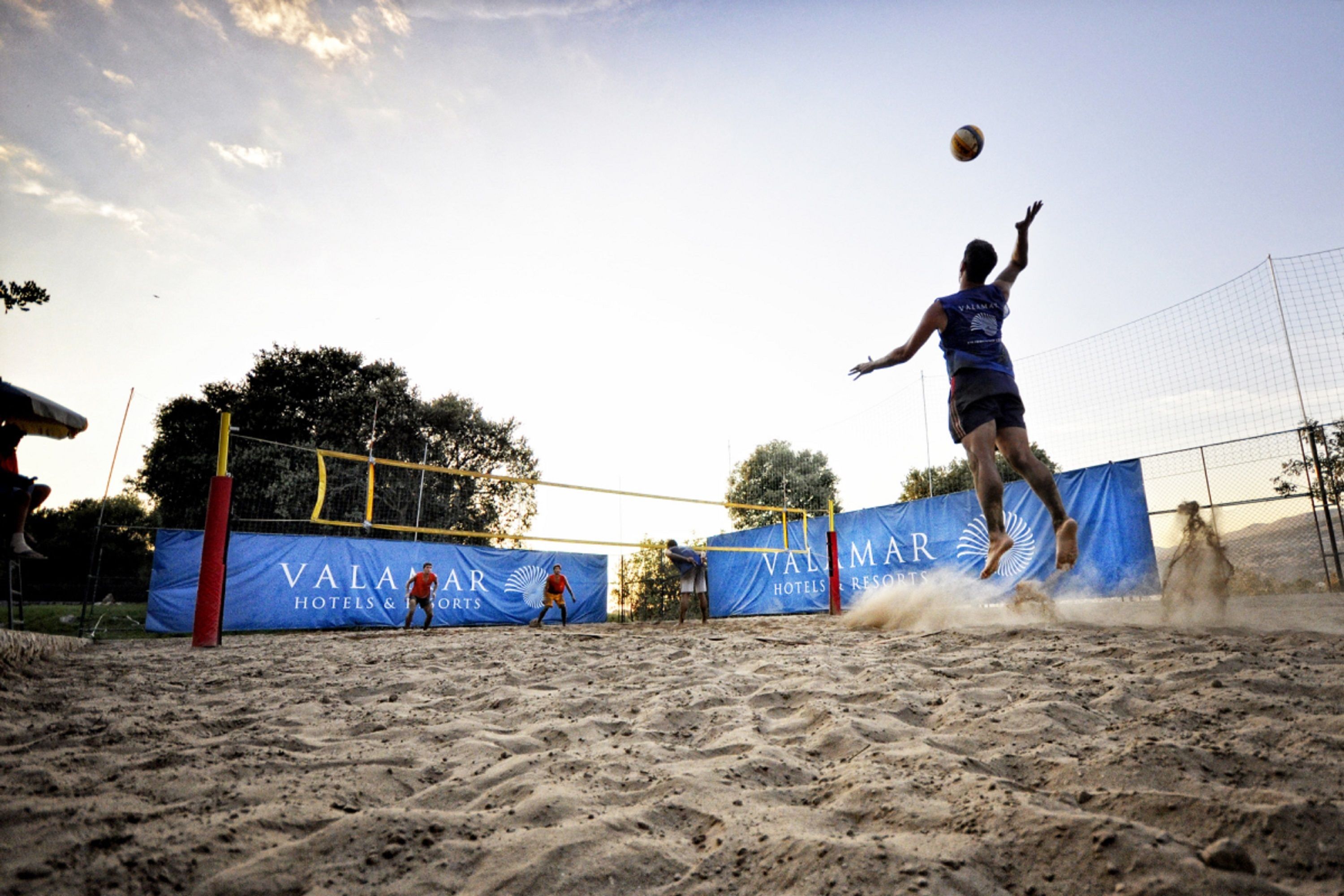 Jogo de voleibol numa quadra de areia, com rede e faixas decorativas ao fundo.