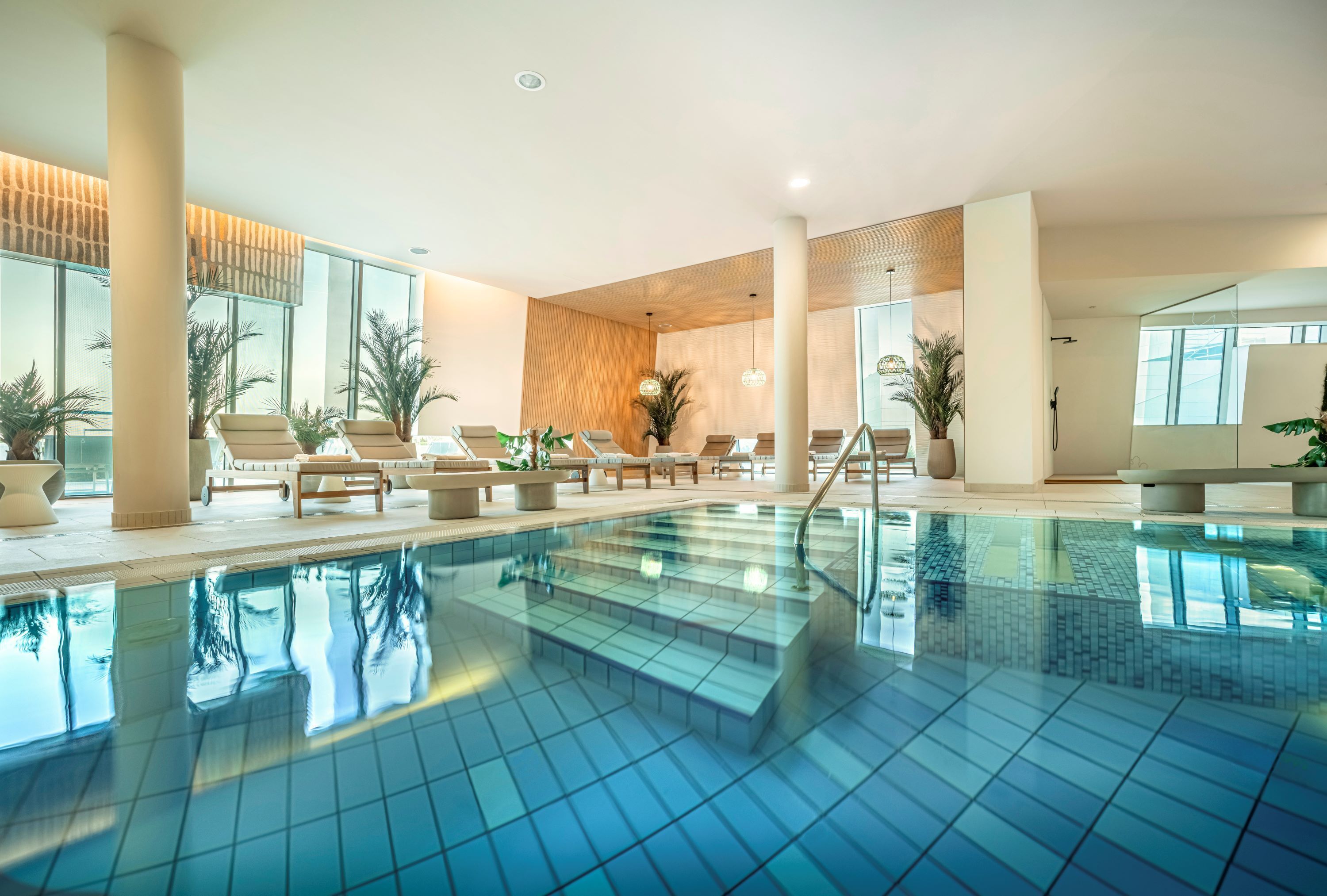 Una piscina interior moderna con baldosas azules cristalinas, sillas de descanso y plantas en macetas.