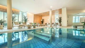 Una piscina interior moderna con baldosas azules cristalinas, sillas de descanso y plantas en macetas.