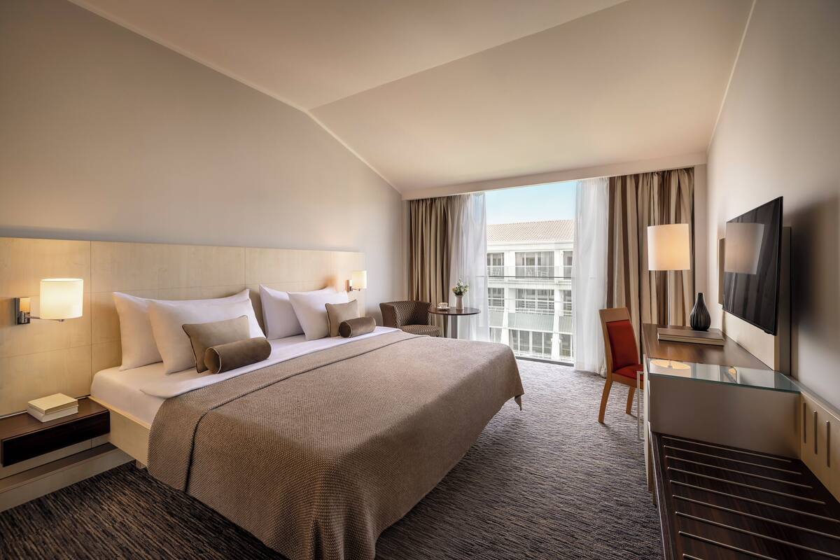 Una habitación de hotel moderna con una cama grande, mesitas de noche, un escritorio con silla, un televisor y un balcón con cortinas.