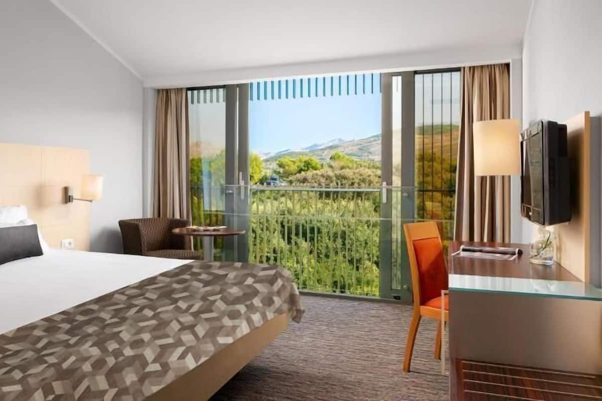 Habitación de hotel con cama, escritorio, silla, televisión y balcón con vistas.