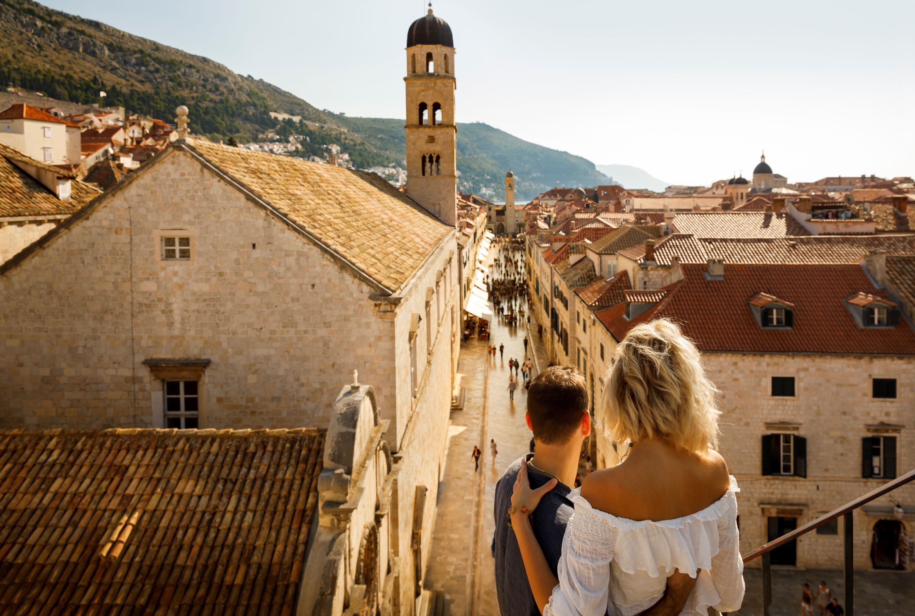 Ein Paar steht auf einer schmalen Gasse mit Blick auf eine historische Stadt mit Steinbauten und einem markanten Turm.