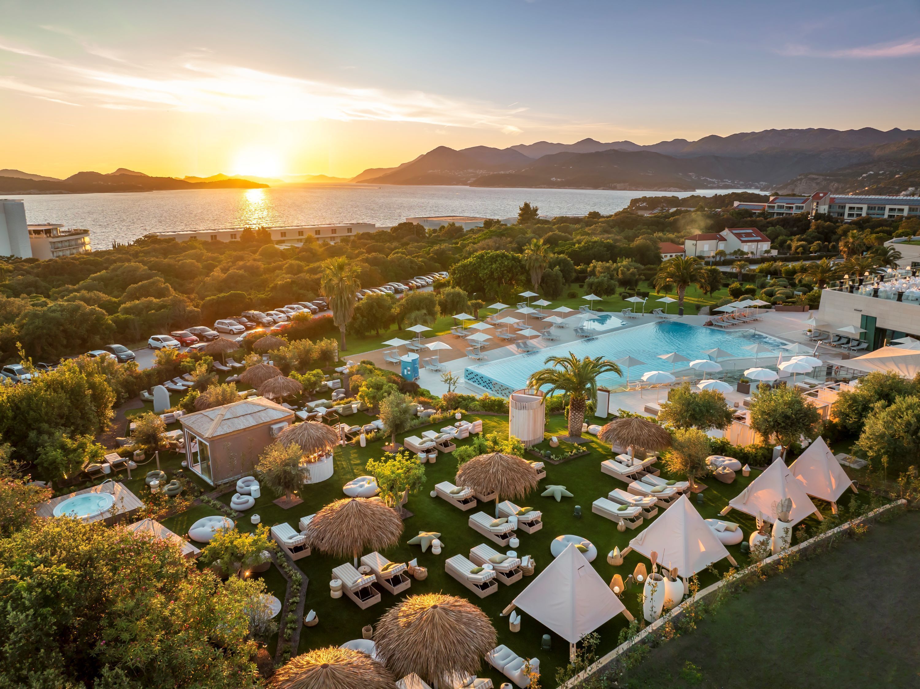 A resort with a large pool, lounge areas, and a view of the sea at sunset.