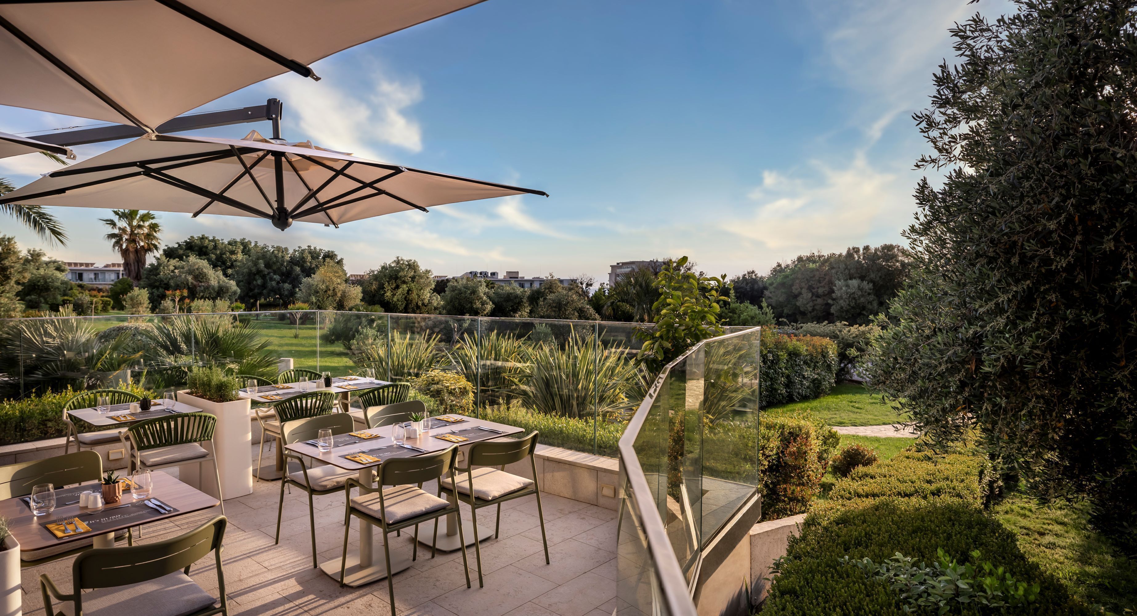 An outdoor dining area with tables and chairs, surrounded by greenery and a clear view of the landscape.