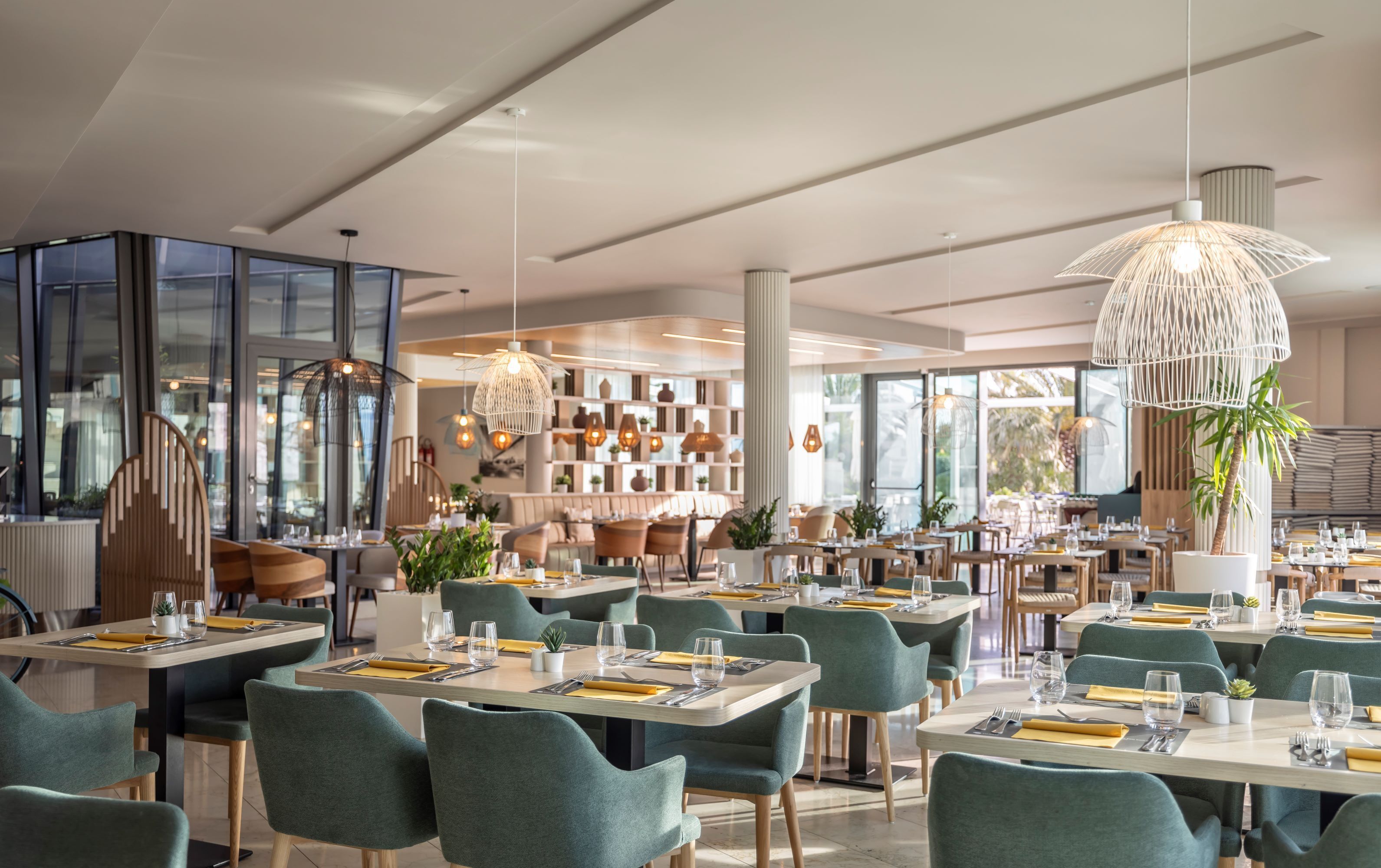 A modern restaurant interior with multiple dining tables, green upholstered chairs, and pendant lighting.