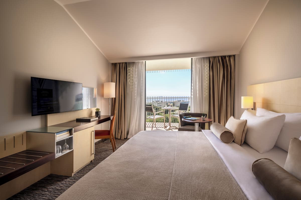 Habitación de hotel con una cama grande, un escritorio con televisor, una pequeña mesa con sillas y un balcón con vistas.