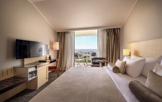 Habitación de hotel con una cama grande, un escritorio con televisor, una pequeña mesa con sillas y un balcón con vistas.