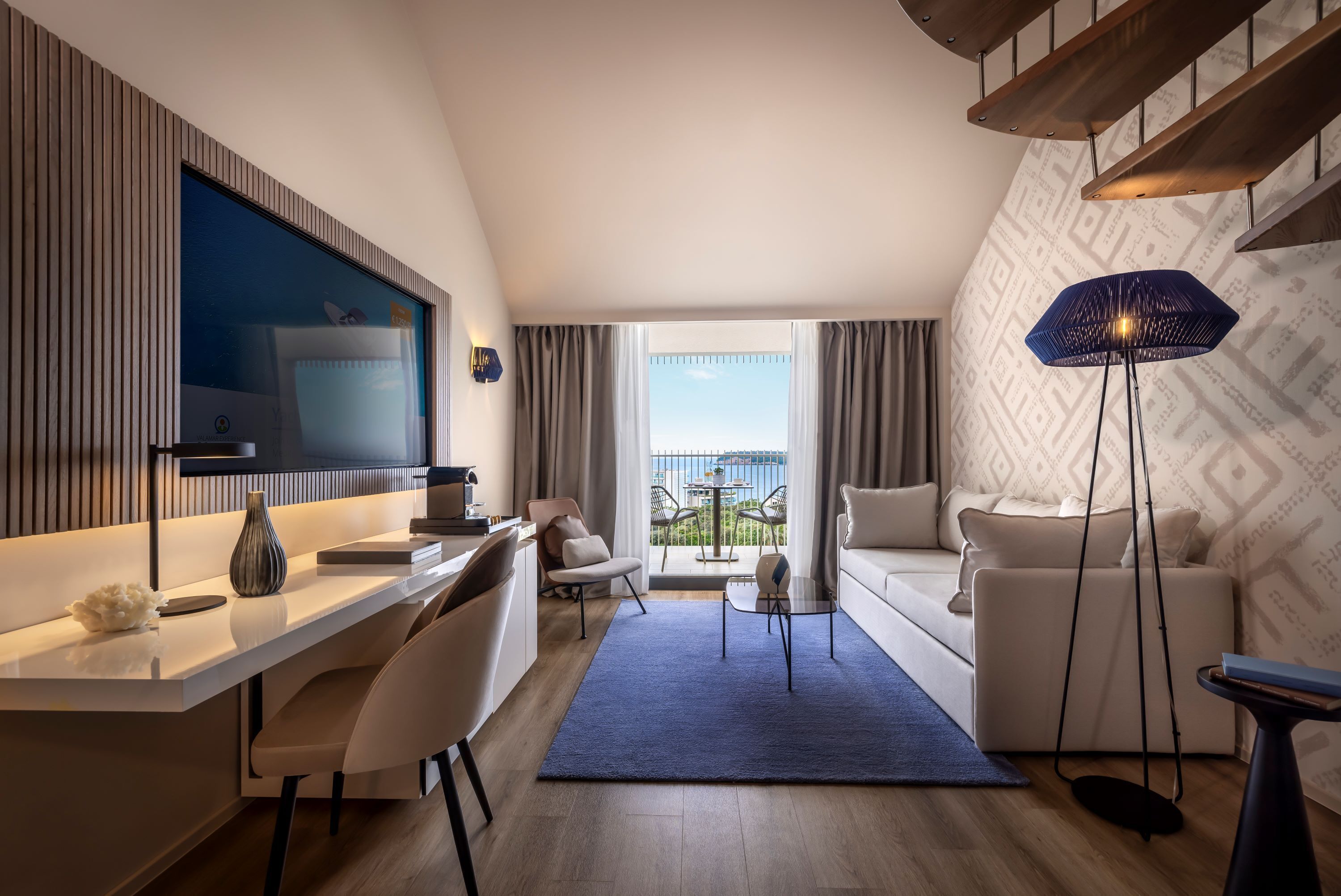 Ein modernes Hotelzimmer mit Schreibtisch, Stuhl, Sofa und einem großen Fenster mit Blick ins Freie.