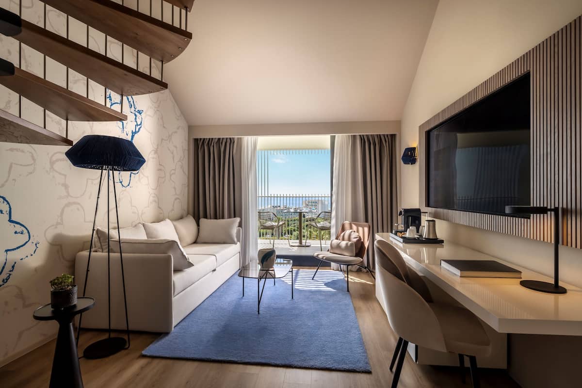 Una sala de estar moderna con sofá, mesa de centro y sillas. Hay un televisor montado en la pared, un balcón con vistas y una escalera.
