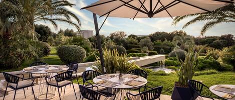 An outdoor patio with round tables and chairs, surrounded by lush greenery and palm trees.