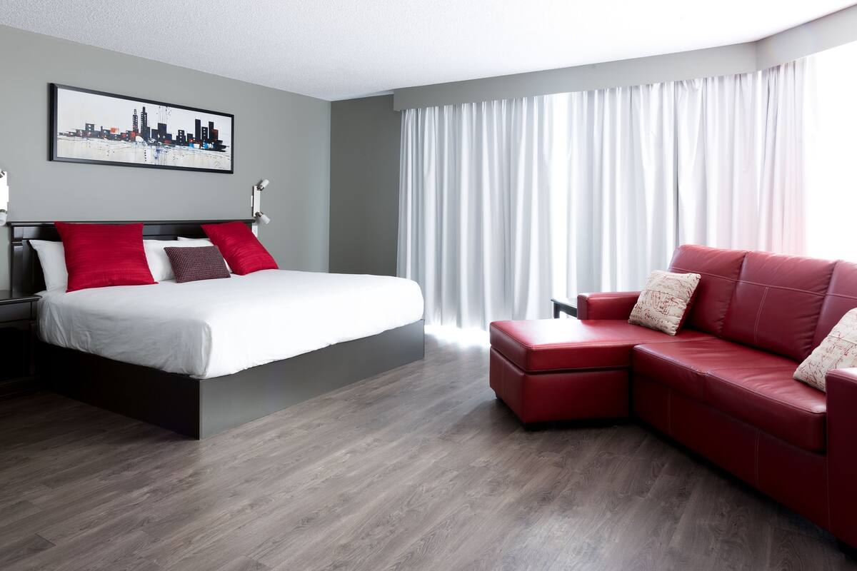 Una habitación de hotel moderna con cama, sofá de cuero rojo, una obra de arte con paisaje urbano en la pared y un ventanal con cortinas.