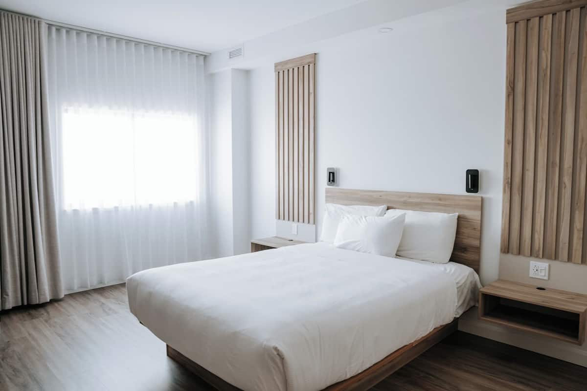 Una habitación de hotel moderna con una cama grande, cabecera de madera, cortinas blancas y suelo de madera.