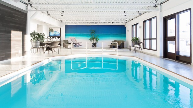 Una piscina interior con vista al océano a través de un ventanal amplio. Hay sillas de descanso y un área de comedor con mesa y sillas.