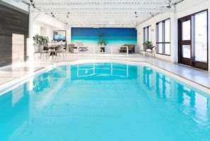 An indoor swimming pool with a view of the ocean through a large window. There are lounge chairs and a dining area with a table and chairs. - Hôtel Universel (Alma)
