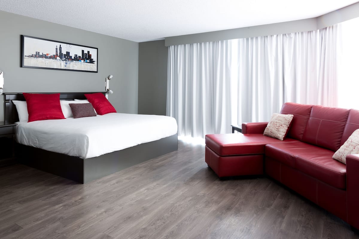Una habitación de hotel moderna con una cama, un sofá de cuero rojo, una obra de arte con un paisaje urbano en la pared y un ventanal con cortinas.