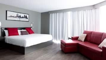 A modern hotel room with a bed, a red leather sofa, a cityscape artwork on the wall, and a large window with curtains.