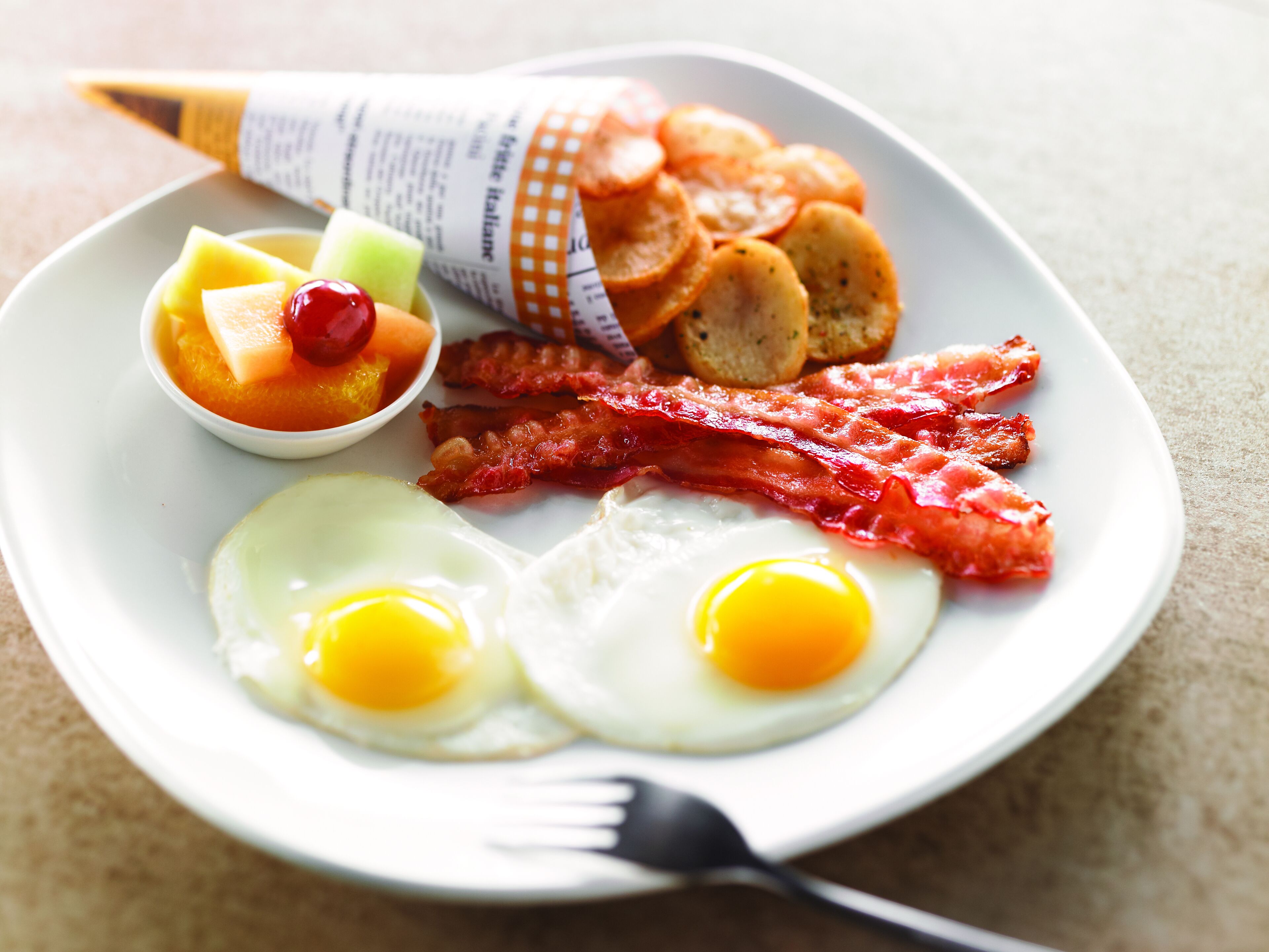 A plate with two fried eggs, bacon, toast, and a small bowl of fruit.