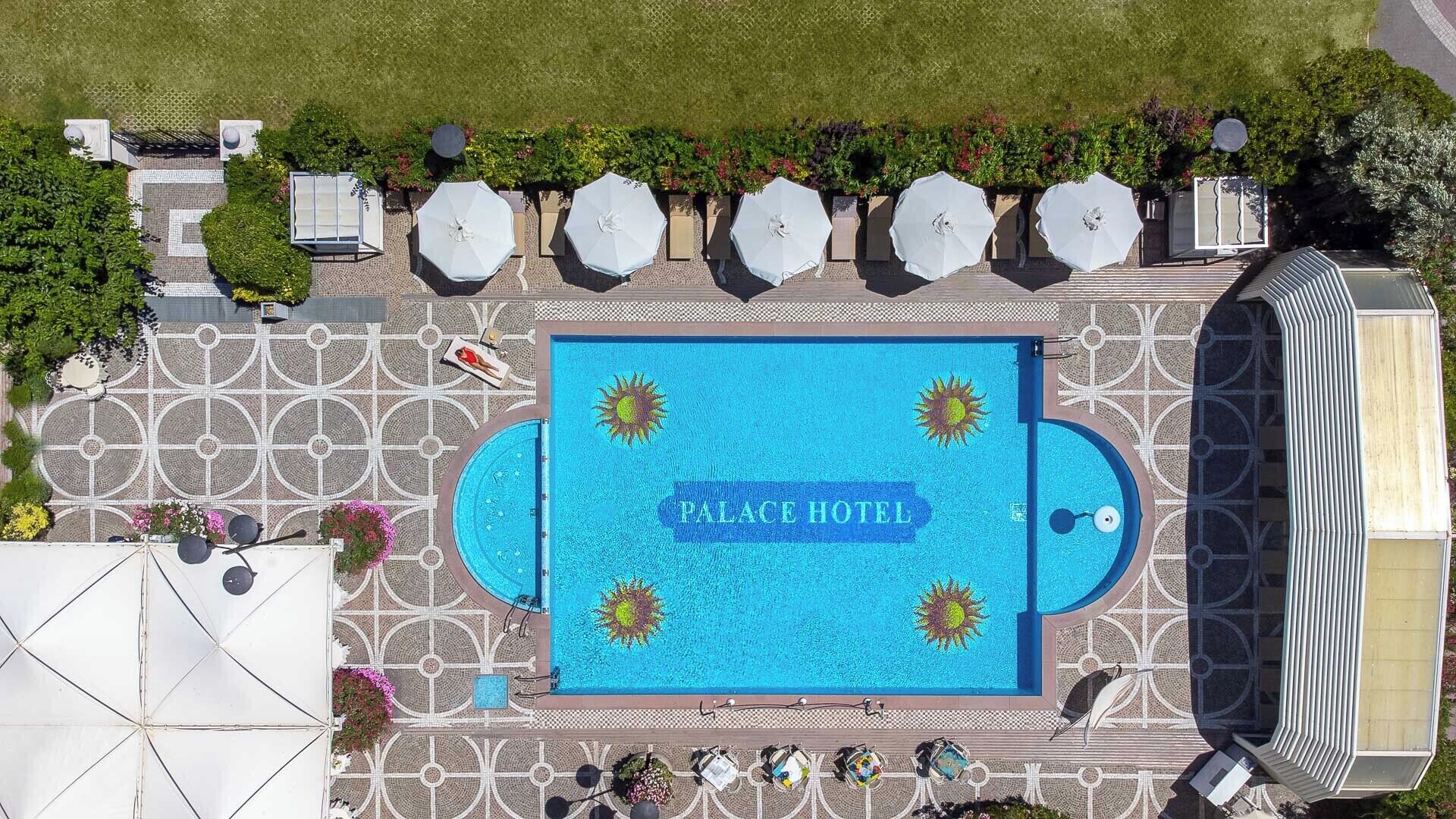 An aerial view of a hotel with a central swimming pool, surrounded by sun-shaped tiles, and multiple umbrella seating areas.