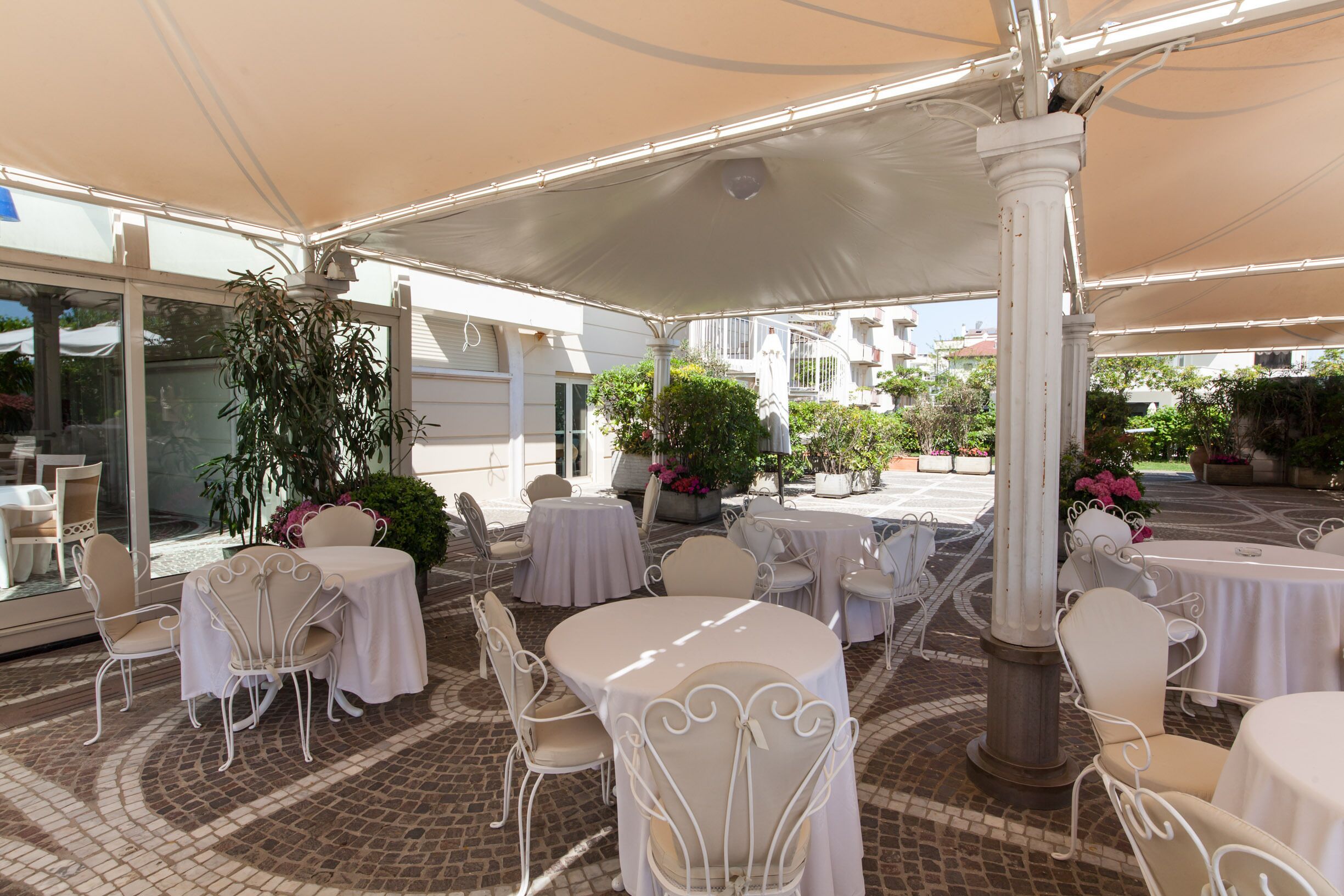 An outdoor patio with round tables and white chairs, covered by a large canopy.