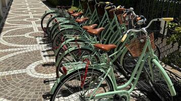 A row of green bicycles with leather seats parked on a patterned pavement, surrounded by greenery and a fence.