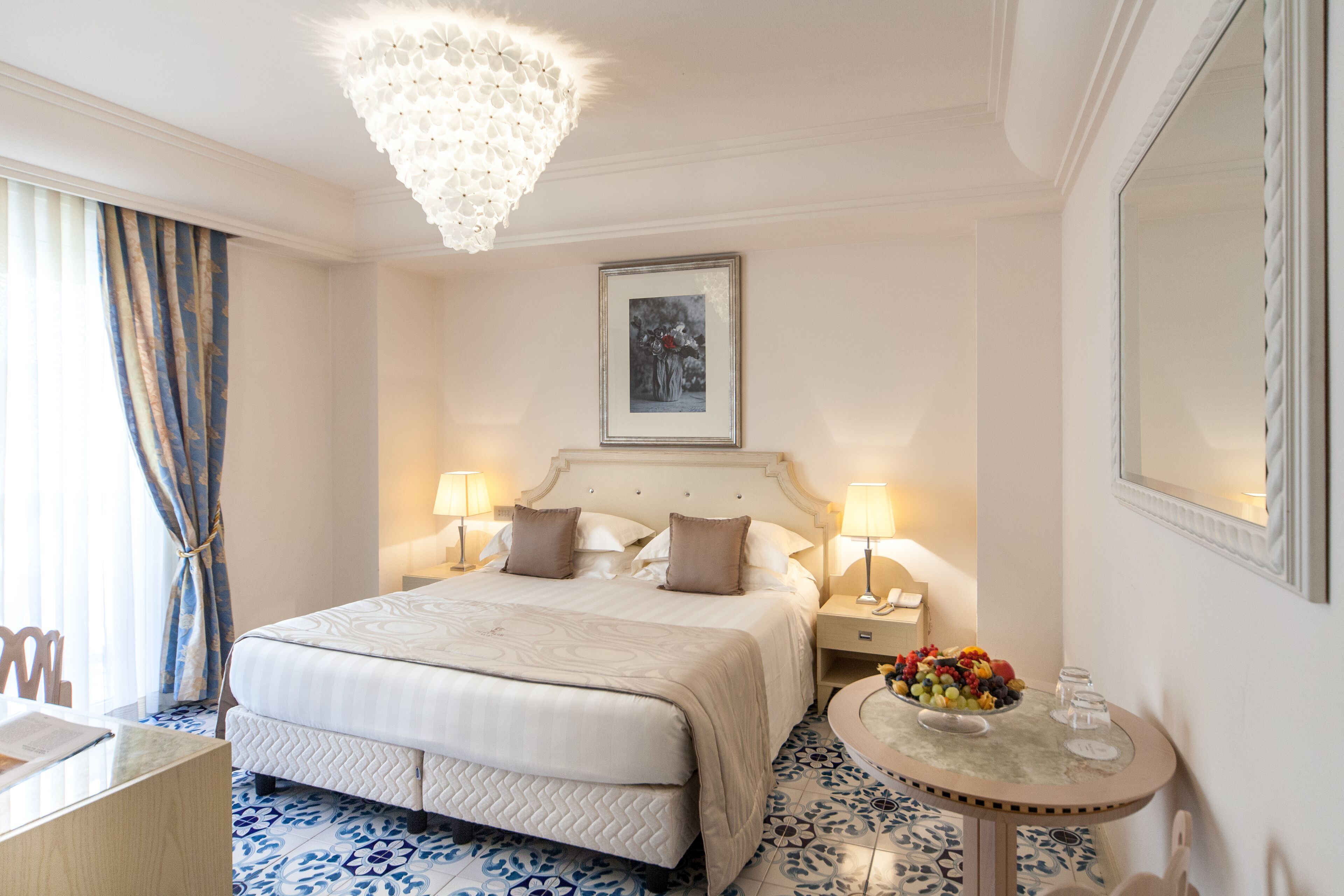 Una habitación de hotel con una cama grande, dos lámparas de noche, una mesa redonda con frutero y una araña.