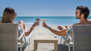 Dos personas brindando con bebidas en una silla de playa junto al océano.