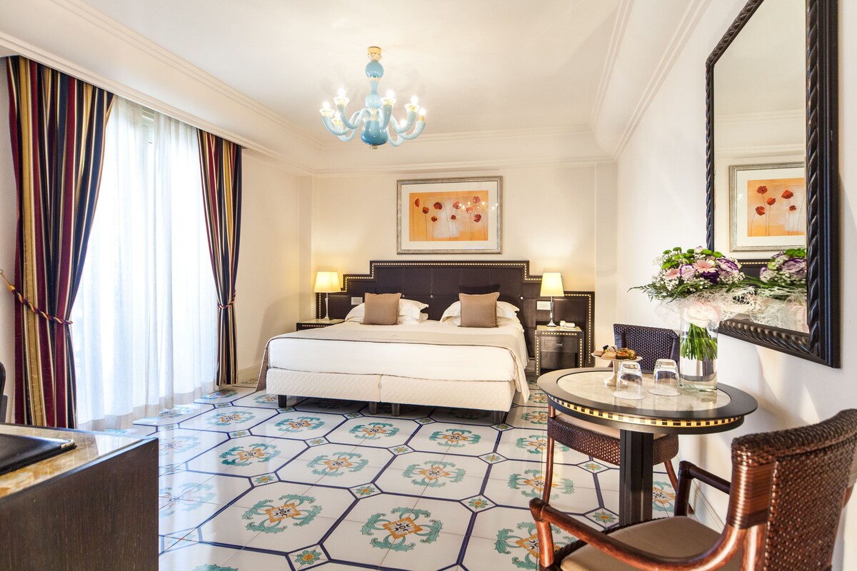 Una camera d'albergo con un letto, comodini, un tavolo rotondo con sedie, un lampadario e opere d'arte alle pareti.