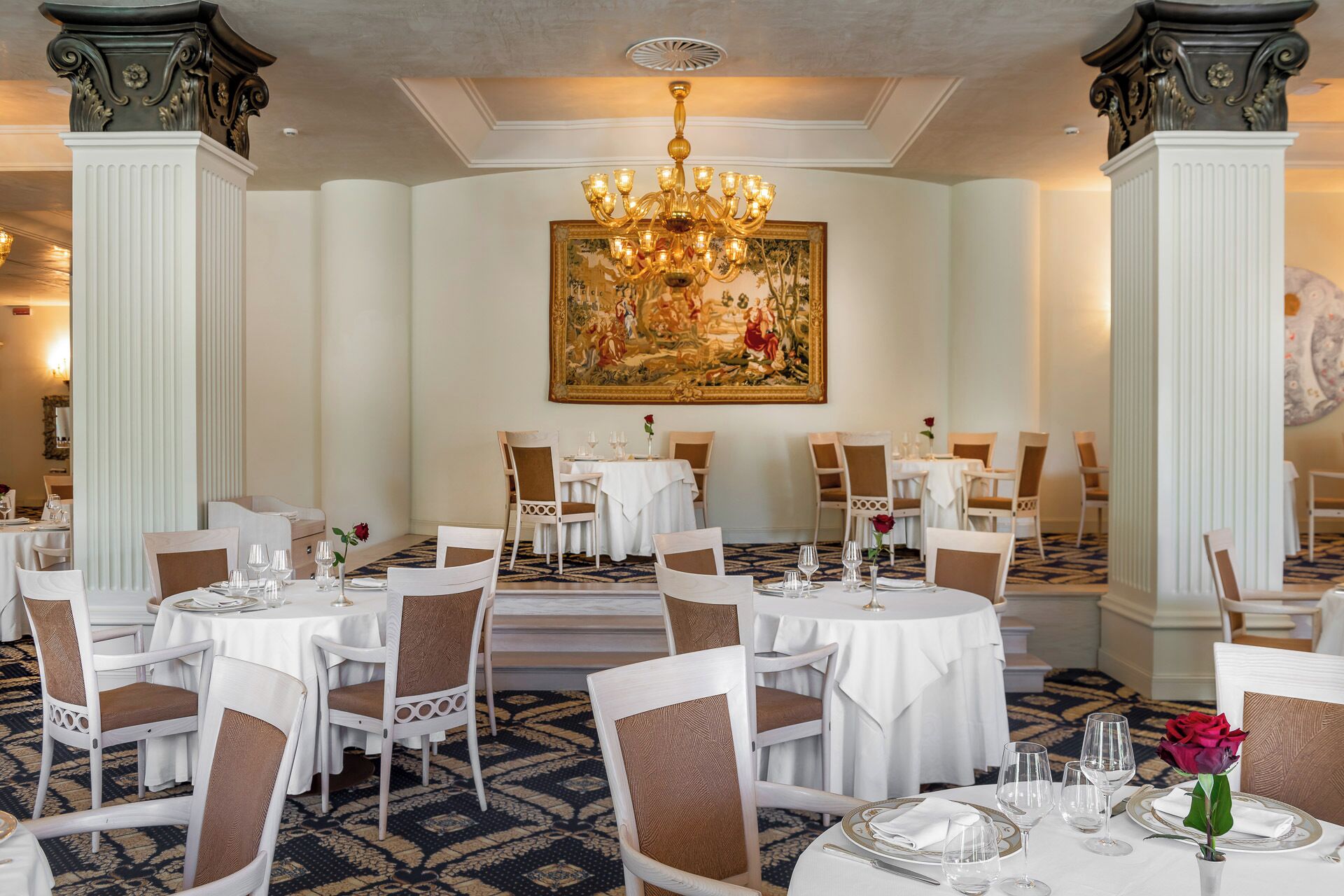 A formal dining room with round tables, white chairs, and a large chandelier.