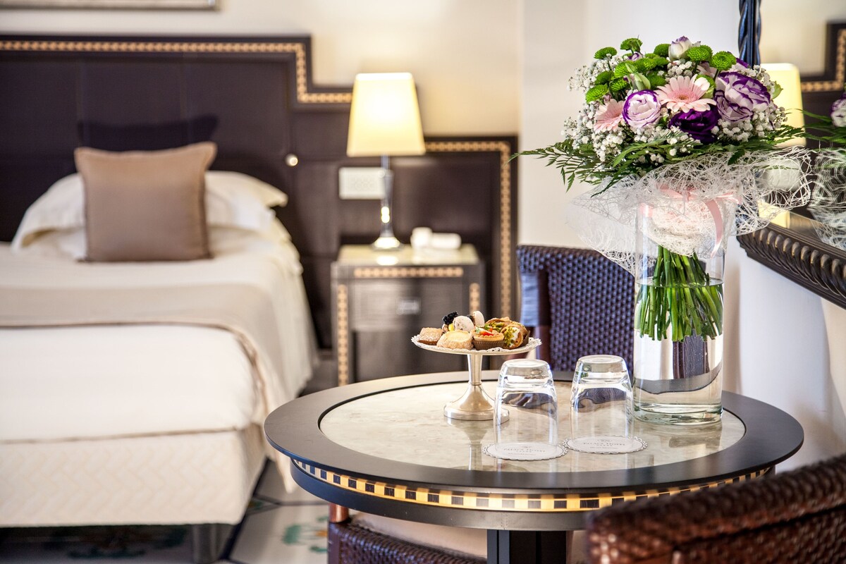 A hotel room with a neatly made bed, a small round table with a vase of flowers, a glass of water, and a plate of pastries.