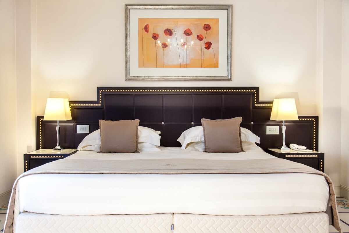 A hotel room with a large bed, two bedside tables with lamps, a framed artwork above the bed, and a headboard with a decorative pattern.