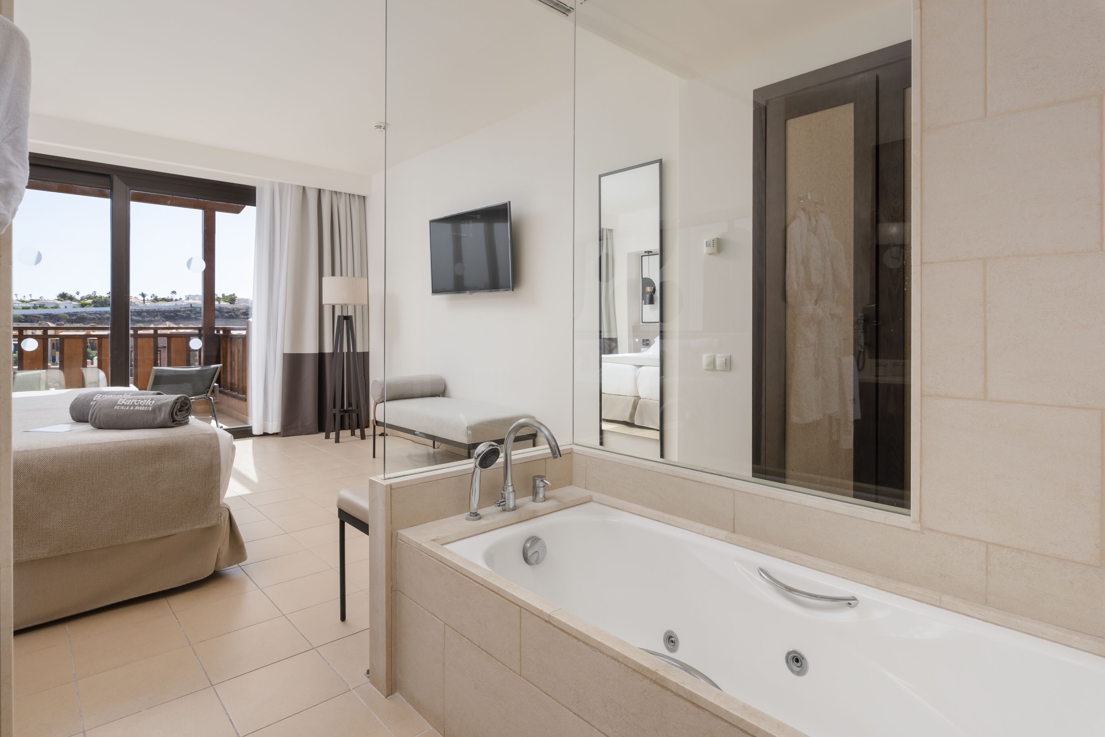 A modern hotel room with a large bathtub, a flat-screen TV, and a balcony with a view of the landscape.