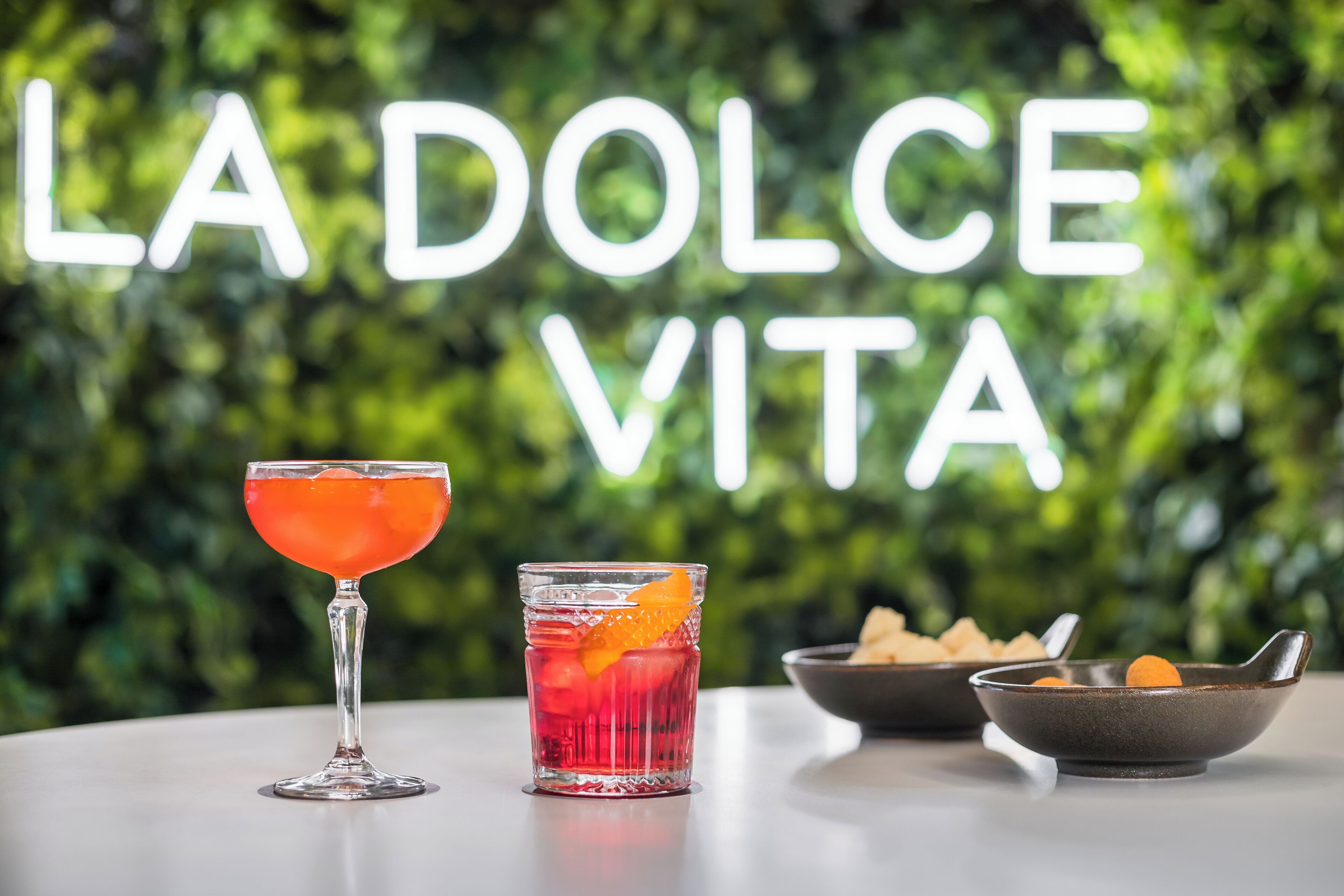 Deux verres de boissons sur une table, avec un arrière-plan verdoyant et feuillu, et le texte « LA DOLCE VITA ».
