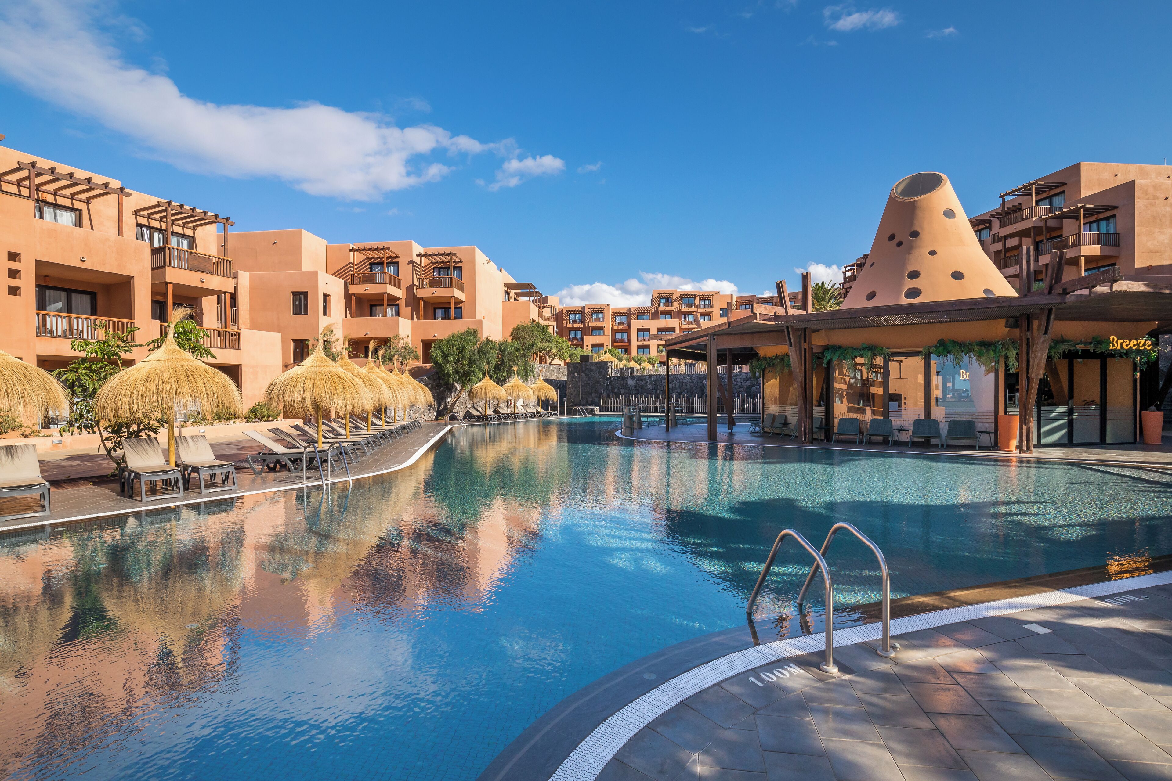 Una piscina con sillas de descanso y sombrillas, rodeada de edificios con balcones y una estructura cónica distintiva.