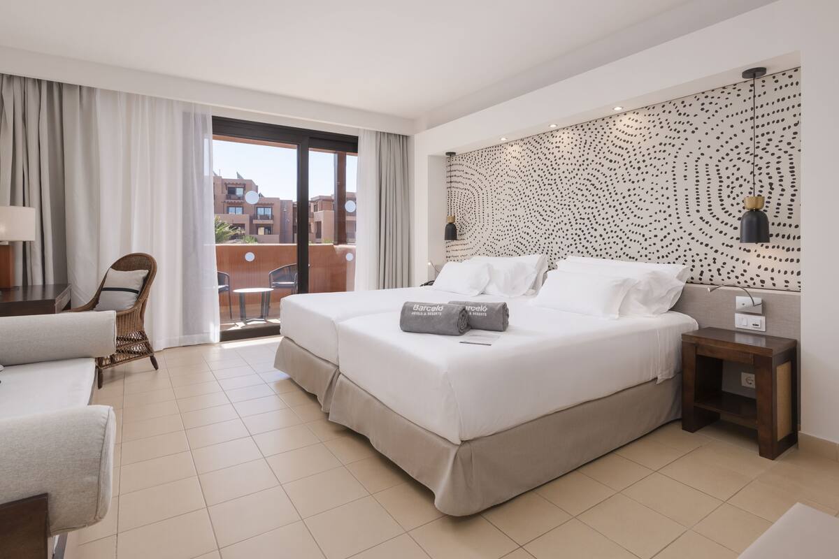 Ein Hotelzimmer mit einem großen Bett, einem Sessel, einem kleinen Tisch und Blick auf die Stadt durch eine Schiebetür.