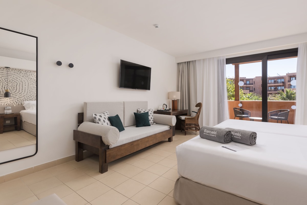 A hotel room with a bed, a sofa, a TV, a desk, and a balcony with chairs.