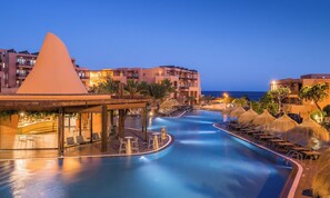 Un espace piscine d’un complexe hôtelier offrant une vue dégagée sur l’océan, plusieurs coins détente avec des parasols, et divers bâtiments en arrière-plan.