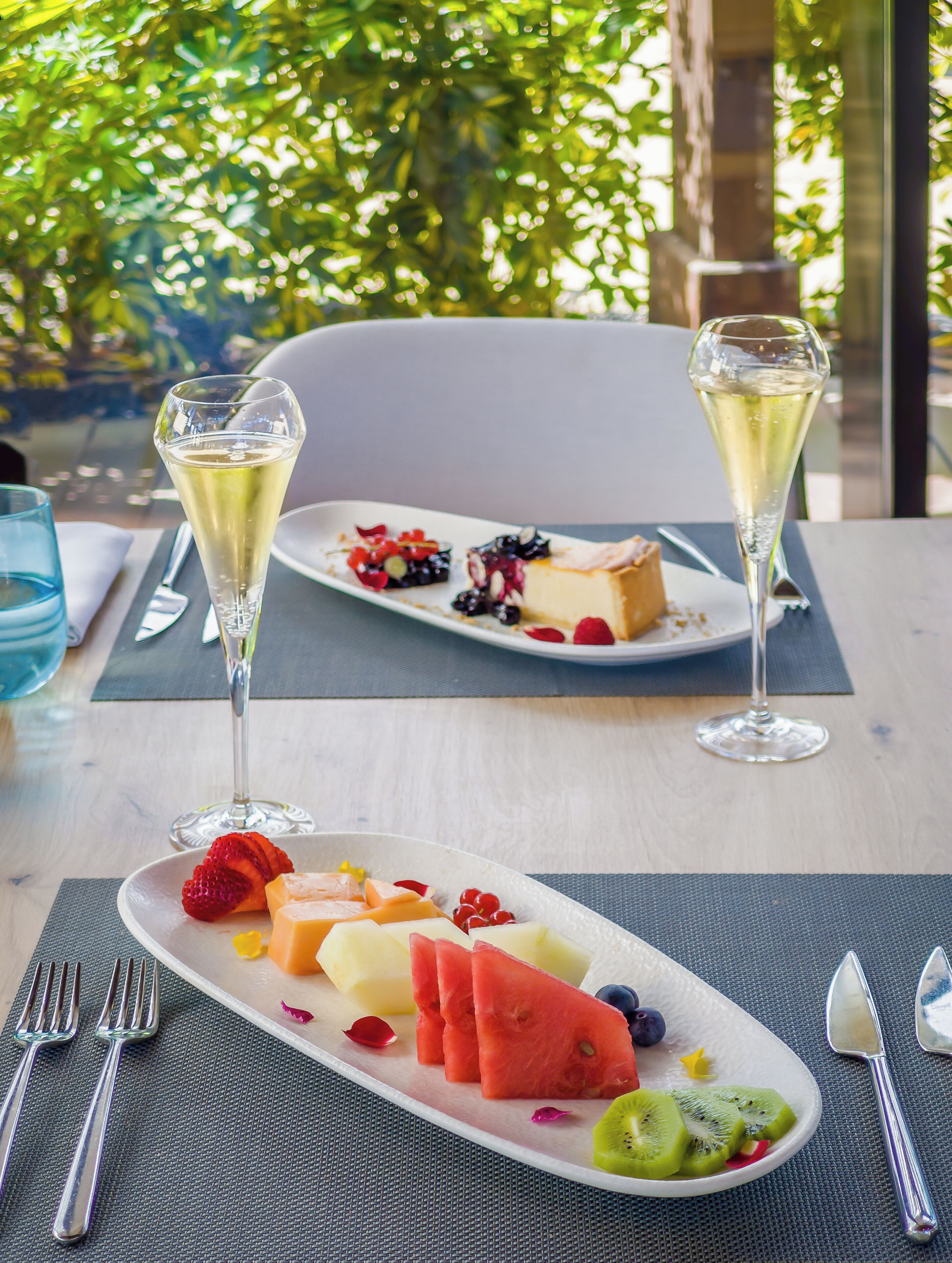 Une table dressée avec une assiette de fruits et deux coupes de champagne, avec en arrière-plan une vue sur la verdure.