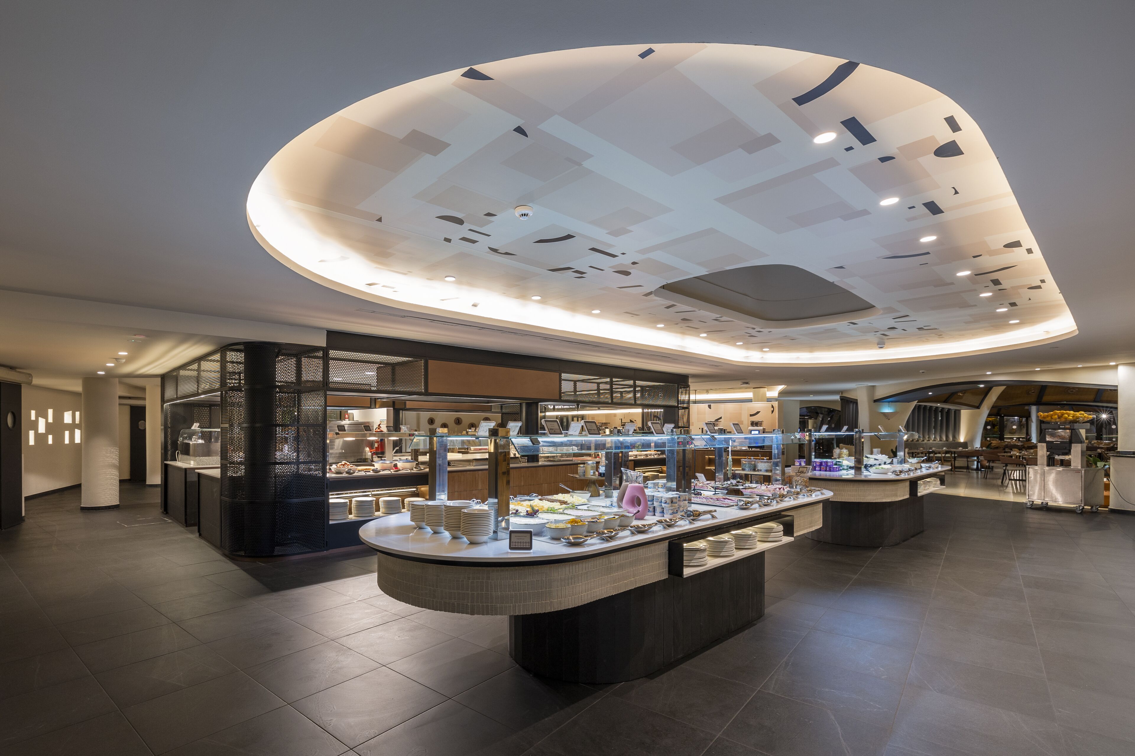 Een moderne hotellobby met een centraal receptiegedeelte, diverse balies met eten en drankjes, en een opvallend plafondontwerp.