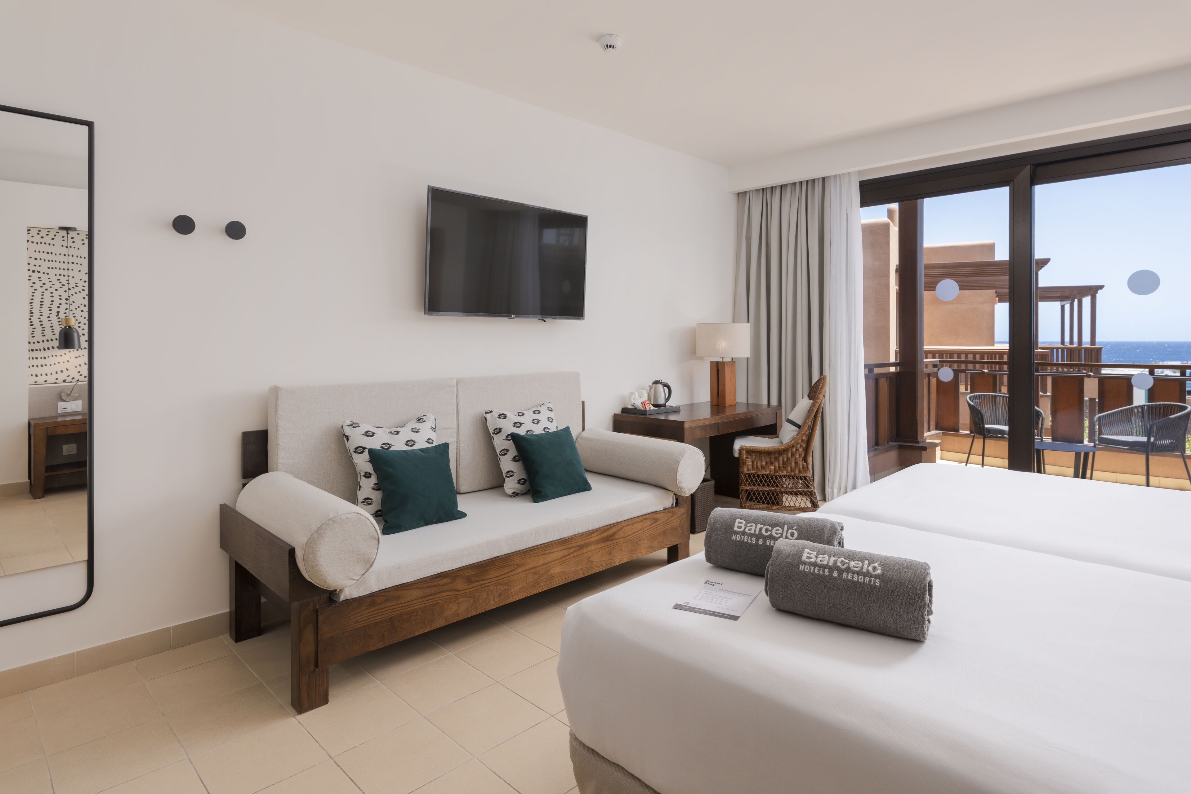 A hotel room with a sofa, a bed, a TV, a balcony with a view of the sea, and a chair.