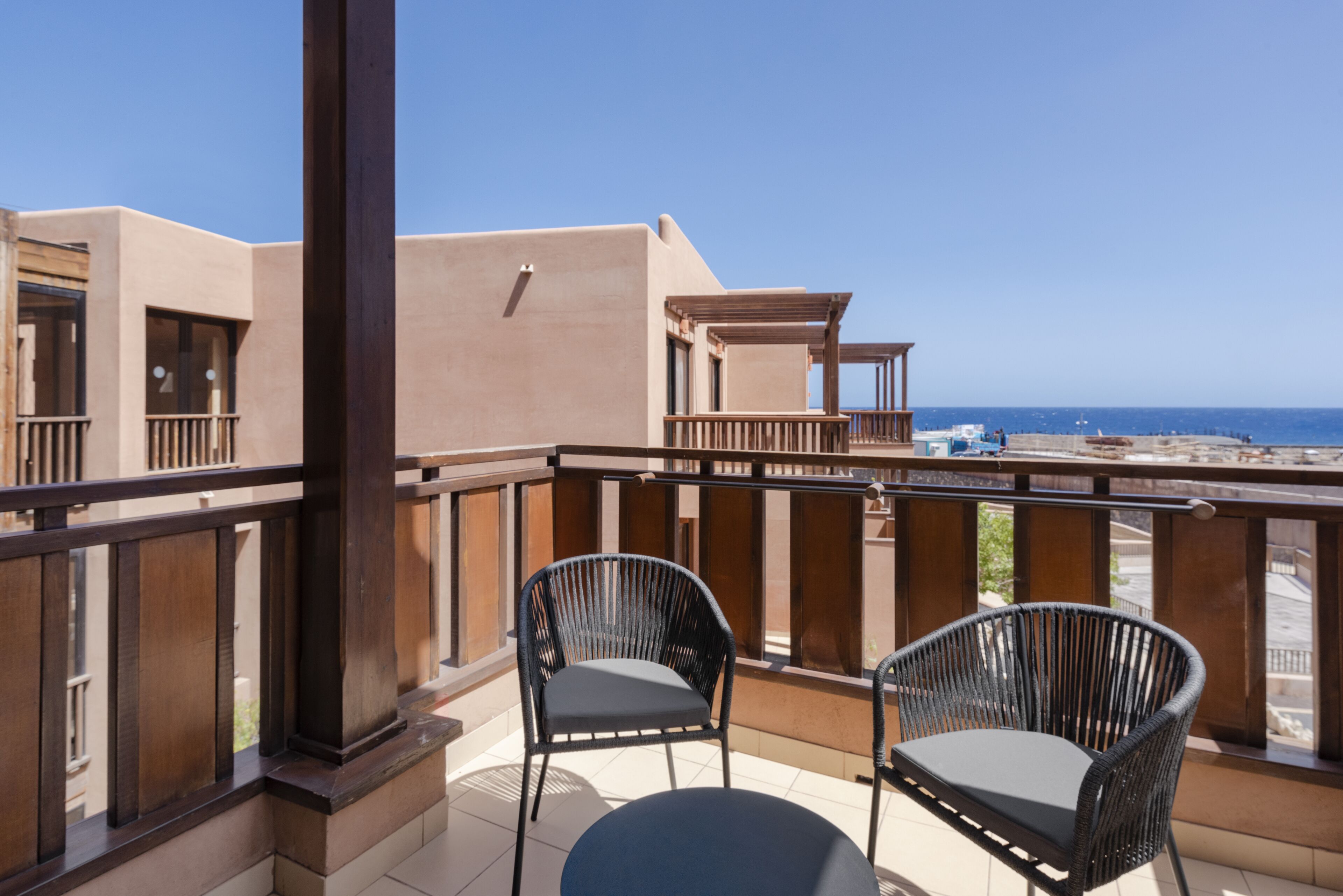 A balcony with two wicker chairs and a table, overlooking a beach and the sea.