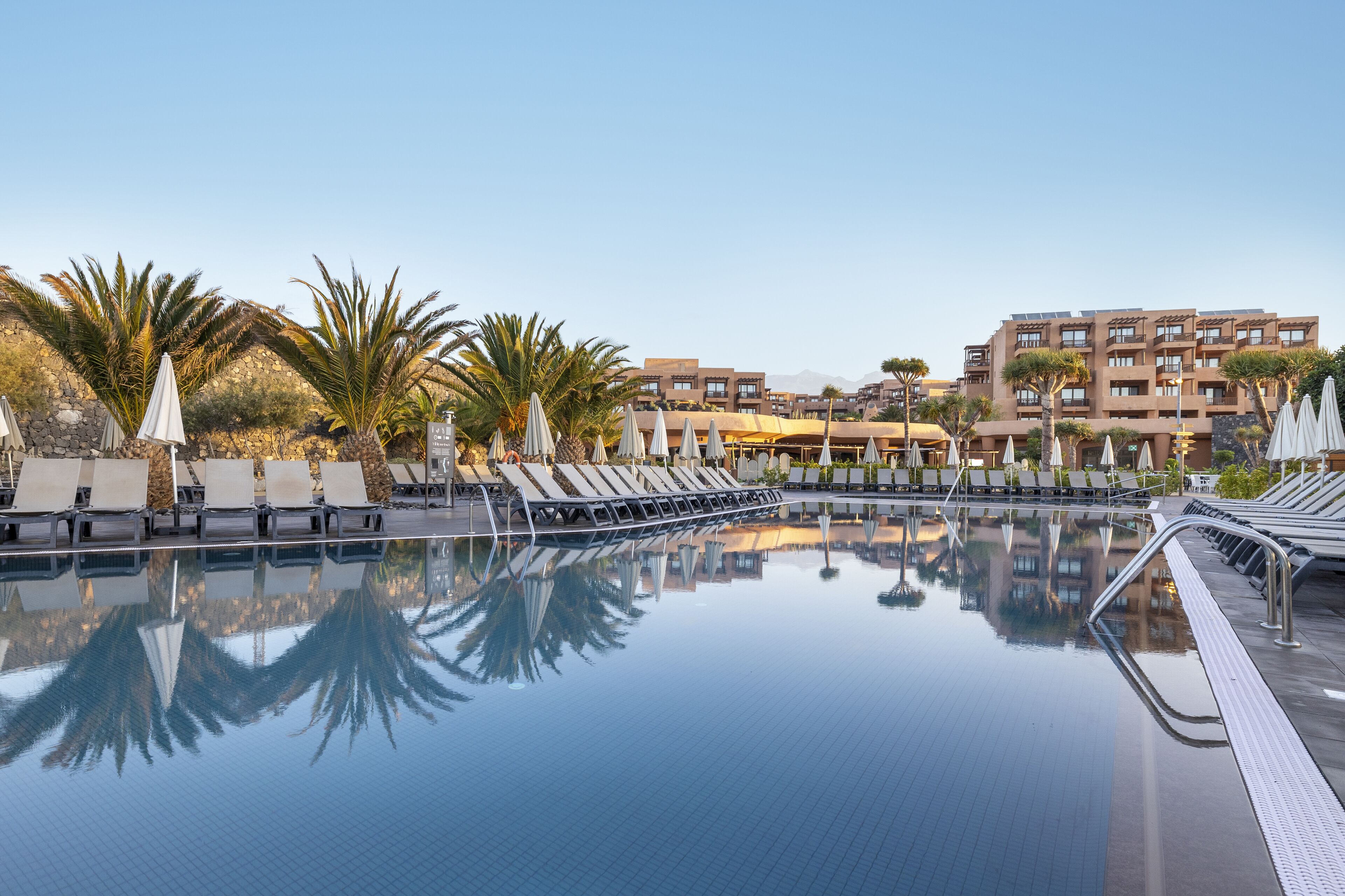 A swimming pool with lounge chairs and umbrellas, surrounded by palm trees and modern buildings.