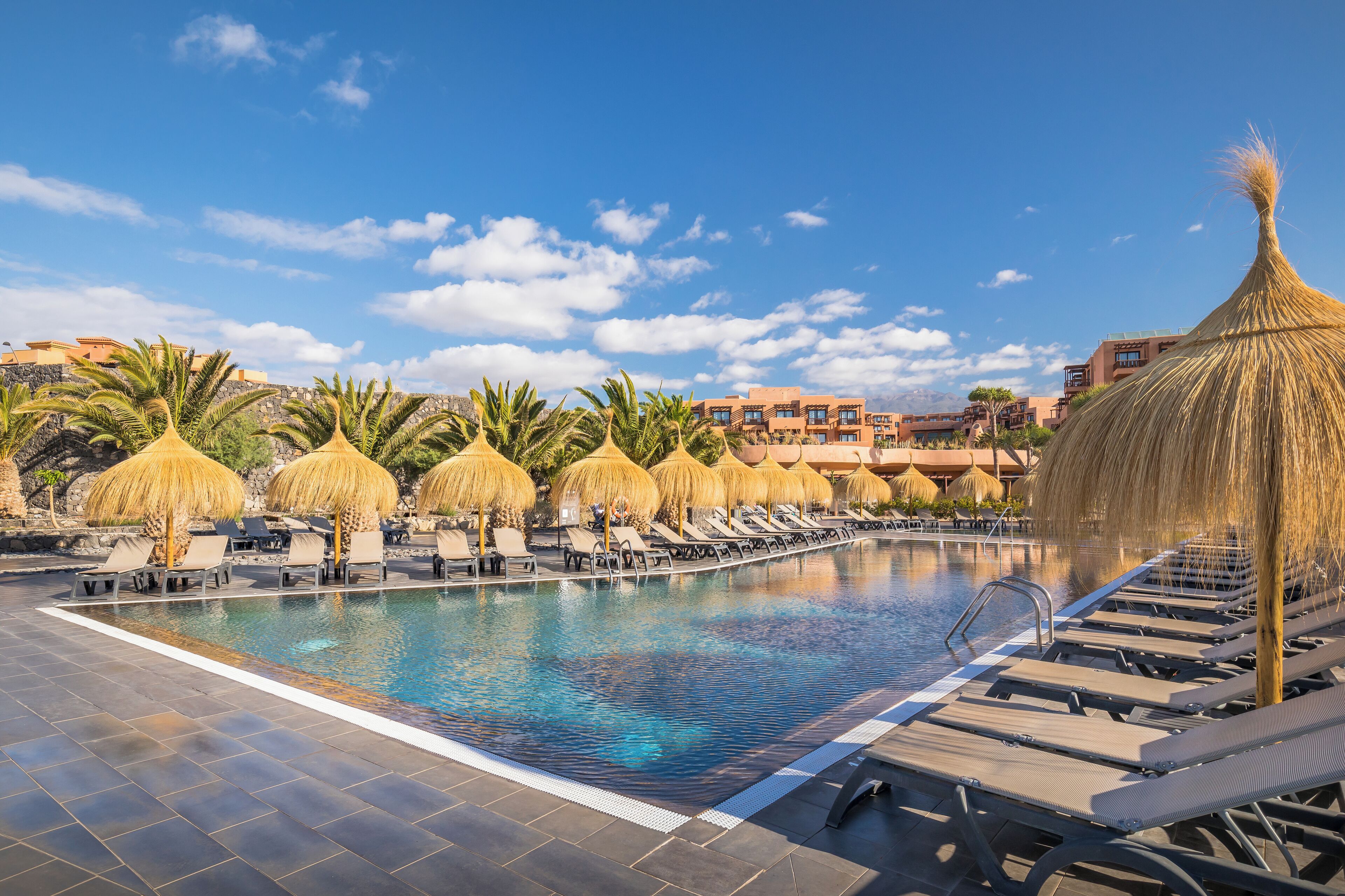 A swimming pool with lounge chairs and thatched umbrellas, surrounded by palm trees and buildings in the background.