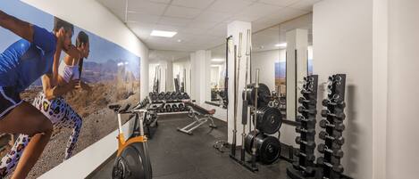 A gym with exercise equipment, a wall with a scenic mural, and a mirror reflecting the equipment.