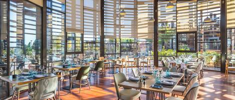 A spacious restaurant with large windows, wooden floors, and modern lighting. Tables are set with chairs and dining essentials.