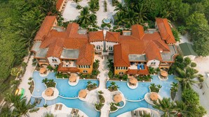 Un resort con varias edificaciones, una zona de piscina central y palmeras alrededor.